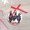 A round wooden ornament featuring a trio of characterful illustrated magpies wearing festive hats. A part of the red ribbon is threaded through the ornament to show where it is hung. A white star is in the top left corner and a couple of candy canes are in the bottom right corner - all sitting on a smoothly textured grey background.