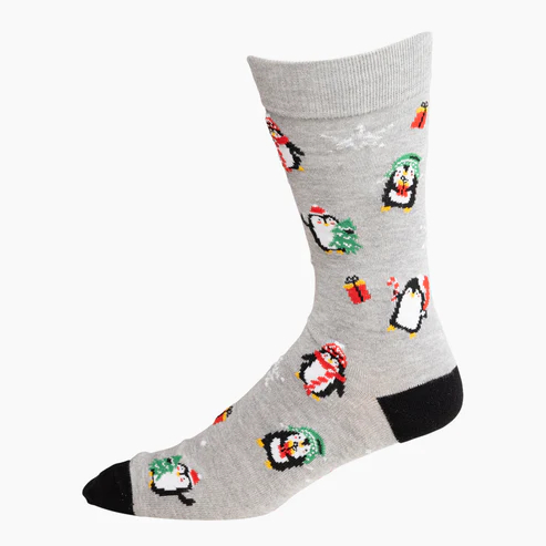 A light grey sock with a black heel and toe, featuring festive cartoon penguins wearing scarves and beanies, holding trees and candy canes.