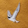 An enamel pin of a Fairy Tern in flight, pinned to a yellow fabric background.