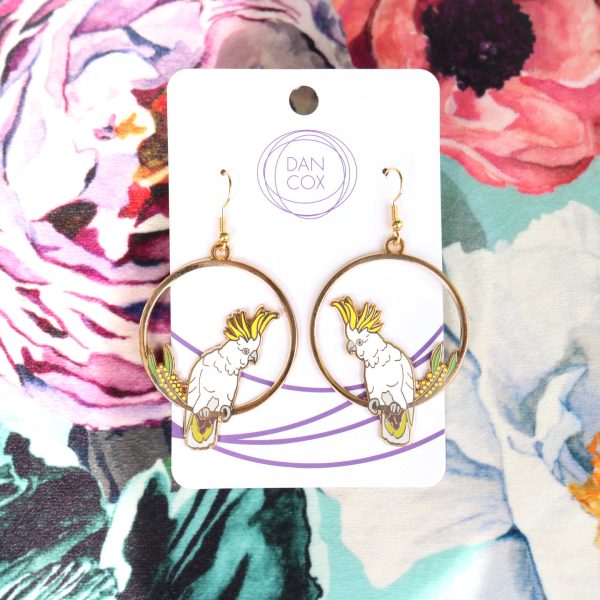 Two circular earrings displayed on a card, sitting on top of a colourful floral background. The earrings feature a Sulphur-crested Cockatoo sitting next to some wattle.